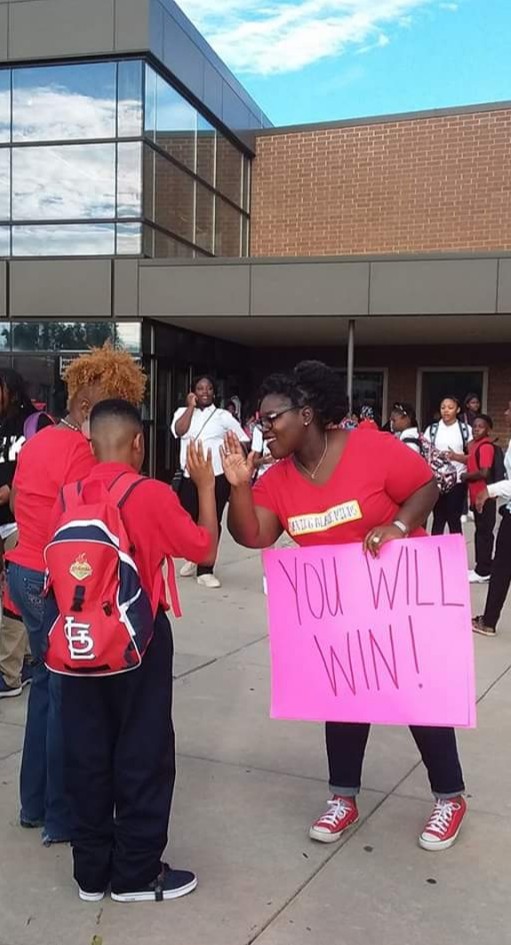A volunteer greets a student at school beside an encouraging sign
